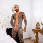 tattooed naked gay male Juna Lucho stroking cock by bed